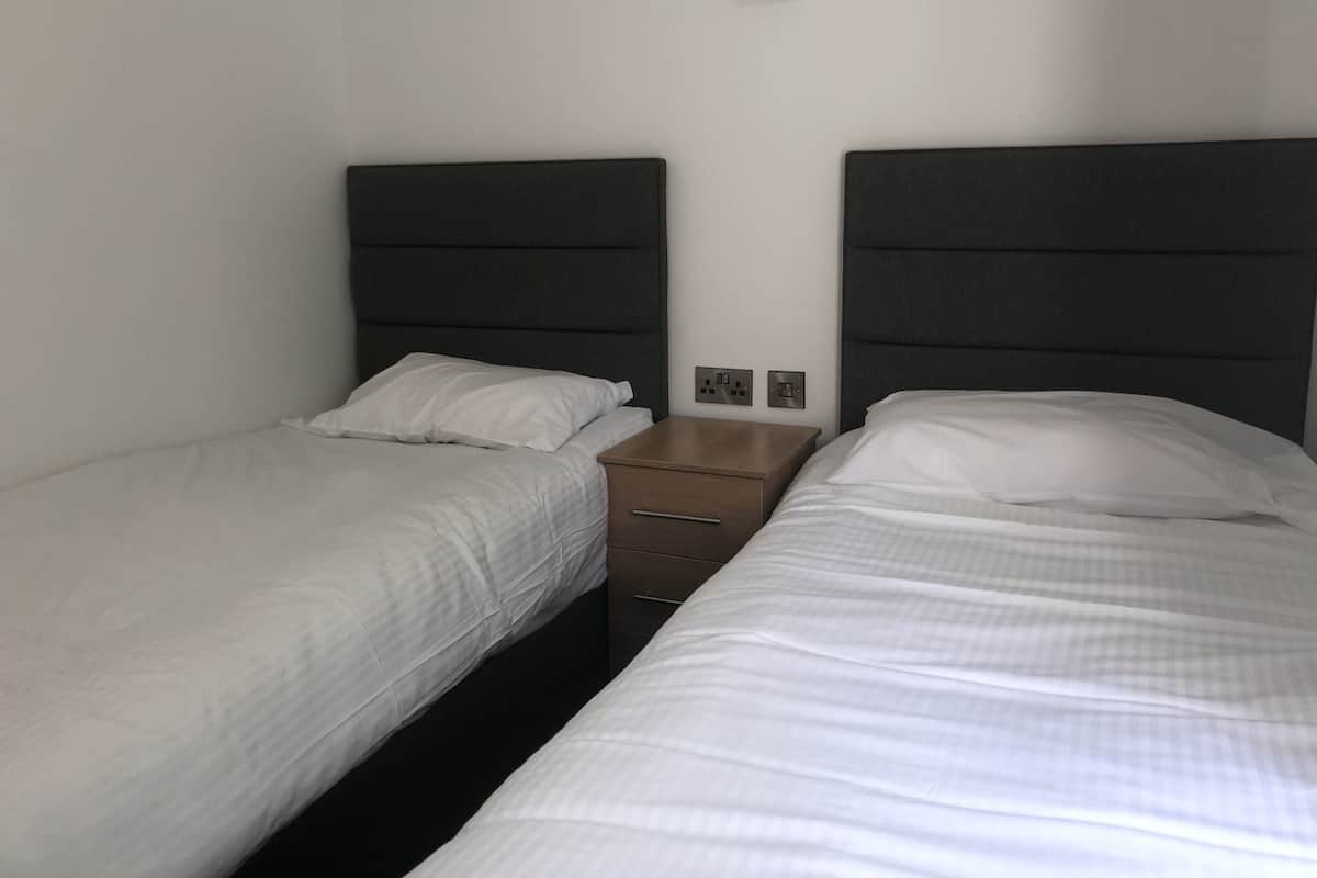 A hotel room with two single beds, each with a dark headboard and white bedding. There is a wooden nightstand between the beds with a power outlet and a small lamp.