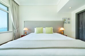 A hotel room with a large bed, two bedside lamps, a window with curtains, and a door. - Maison Privee - Beach Access Apt w/ Burj & Sea Views on The Palm (Dubai)