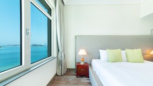 A hotel room with a large window, a bed with white linens and green pillows, a nightstand with a lamp, and a wooden floor. - Maison Privee - Beach Access Apt w/ Burj & Sea Views on The Palm (Dubai)