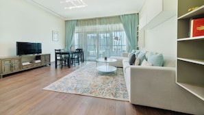 A living room with a sofa, a coffee table, a TV, a dining table with chairs, and a bookshelf. - Maison Privee - Beach Access Apt w/ Burj & Sea Views on The Palm (Dubai)