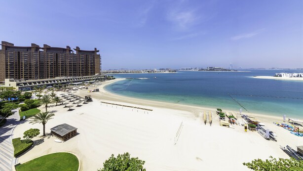 A beachfront hotel with a large building, a sandy beach, and a clear blue sea. - Maison Privee - Beach Access Apt w/ Burj & Sea Views on The Palm (Dubai)
