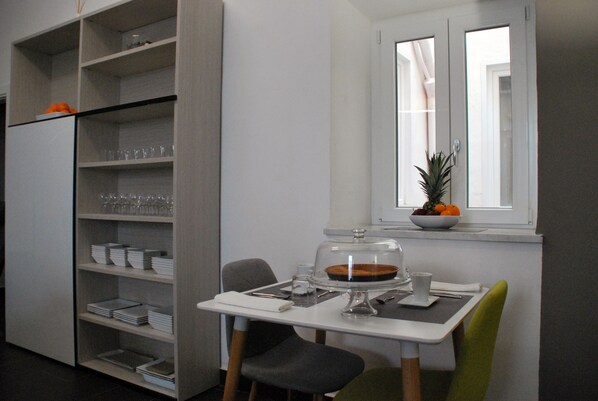 A compact kitchen with a dining area, including a table with chairs, a window with a view, and a built-in shelf with dishes and glasses.