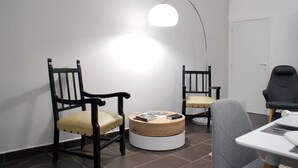 Een kamer met twee houten stoelen met beklede zittingen, een ronde houten tafel met een witte poot en een moderne vloerlamp.