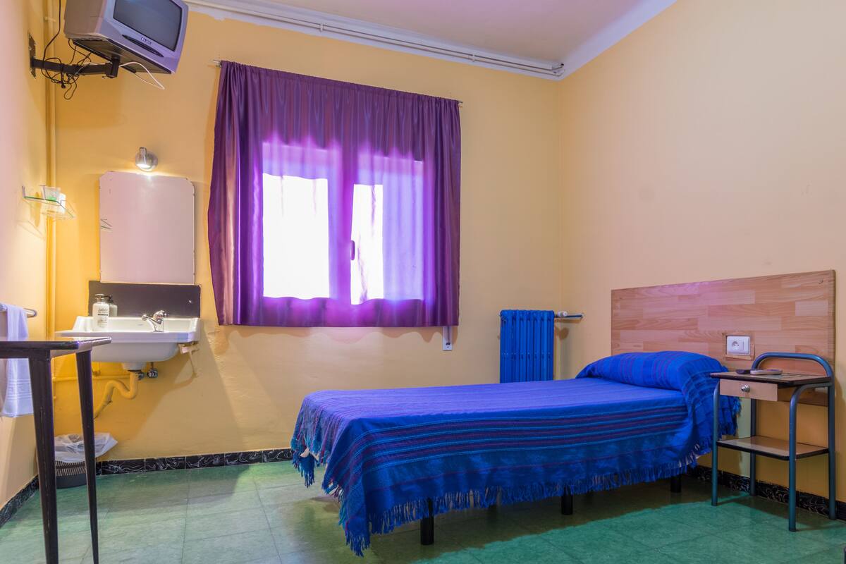 A small room with a bed, a sink, a window with purple curtains, and a television mounted on the wall.