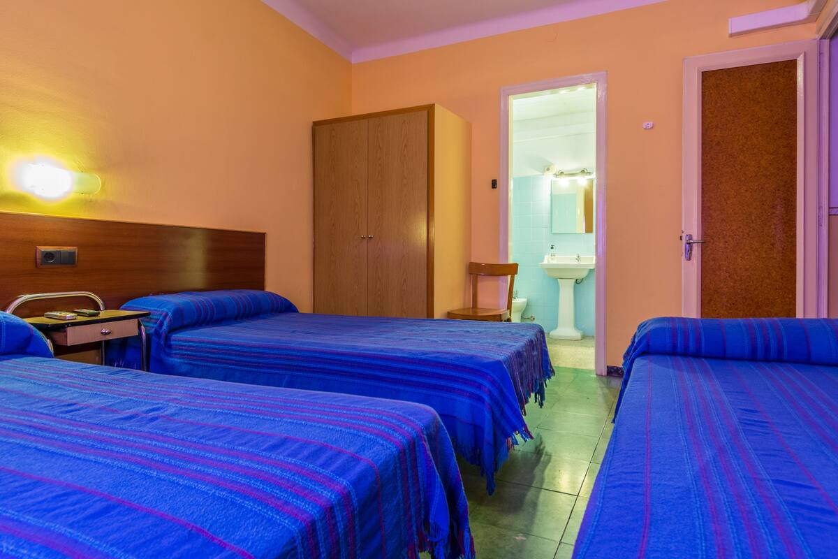 A hotel room with two beds, a wooden wardrobe, a small table with a lamp, and a bathroom visible through an open door.