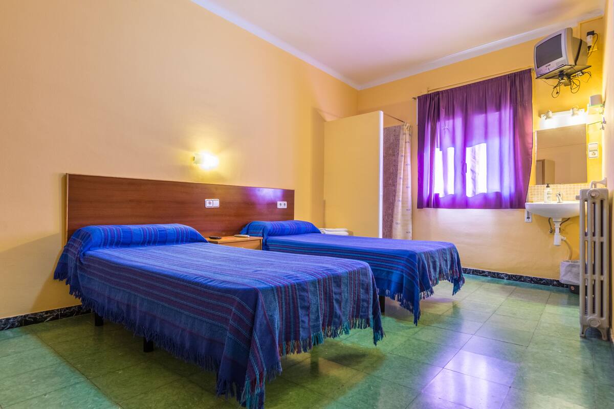 A hotel room with two beds, a wooden headboard, a bathroom with a mirror and sink, and a window with purple curtains.