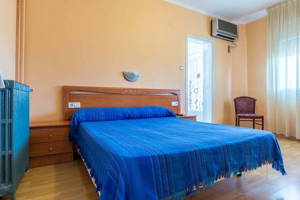 A bedroom with a wooden bed, a nightstand, a chair, and an air conditioning unit on the wall.