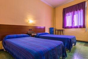 Habitación de hotel con dos camas, cabecera de madera, mesita de noche, una silla y una ventana con cortinas moradas.