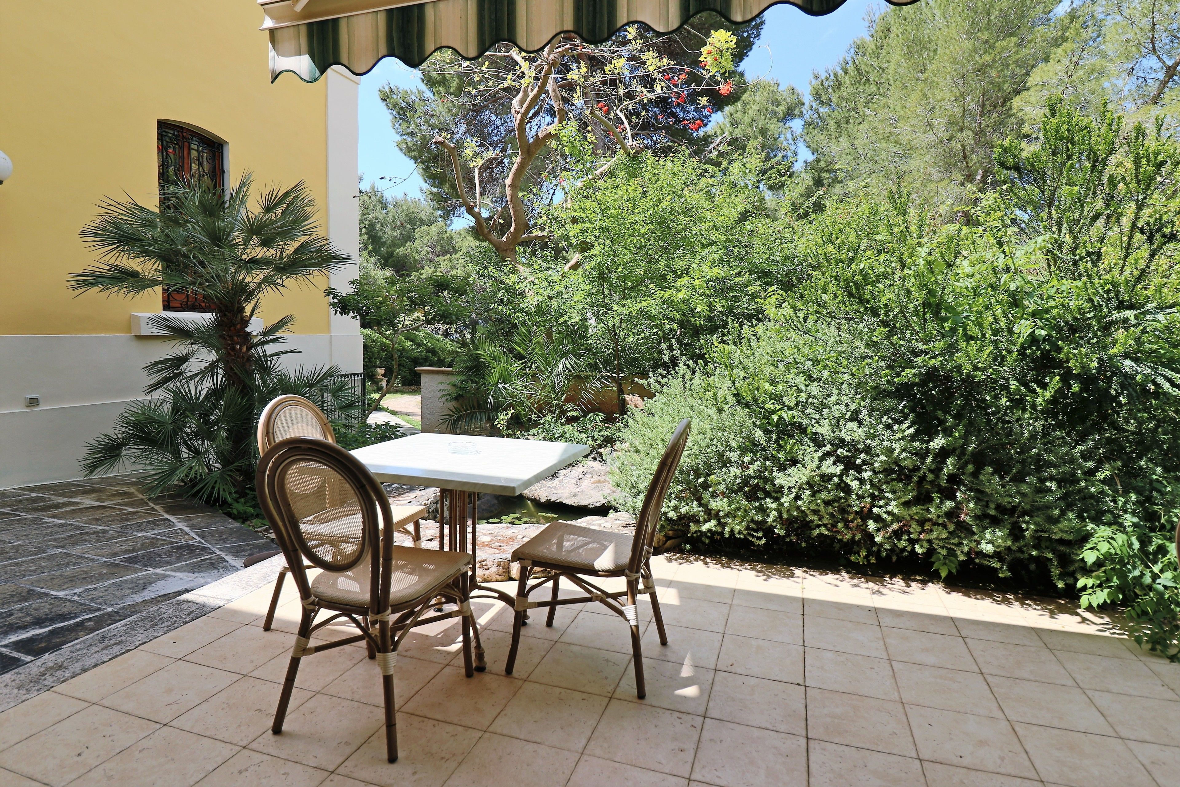 An outdoor patio with a table and chairs, surrounded by lush greenery and a yellow building with a window.