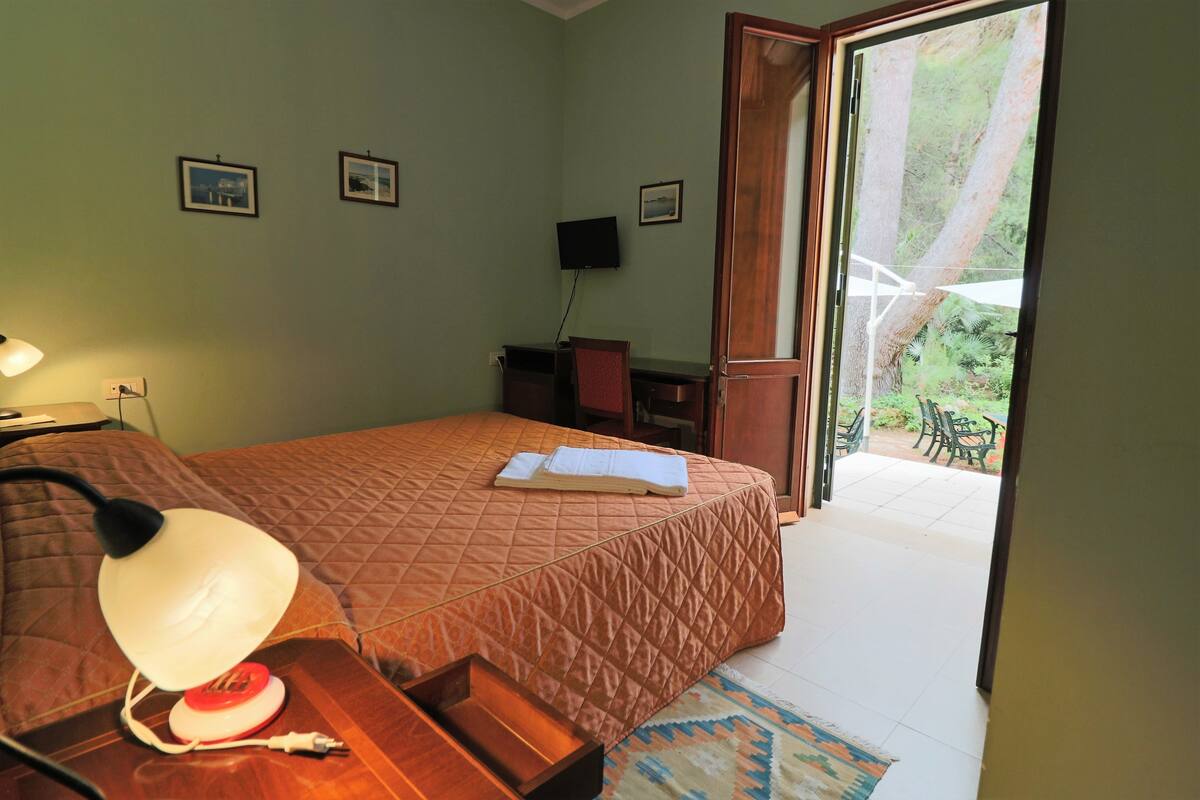 Quarto com cama, mesa de cabeceira, abajur e vista para um jardim por uma porta aberta.