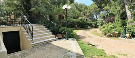 A stone staircase leading to a beige door, surrounded by lush greenery and a paved path.