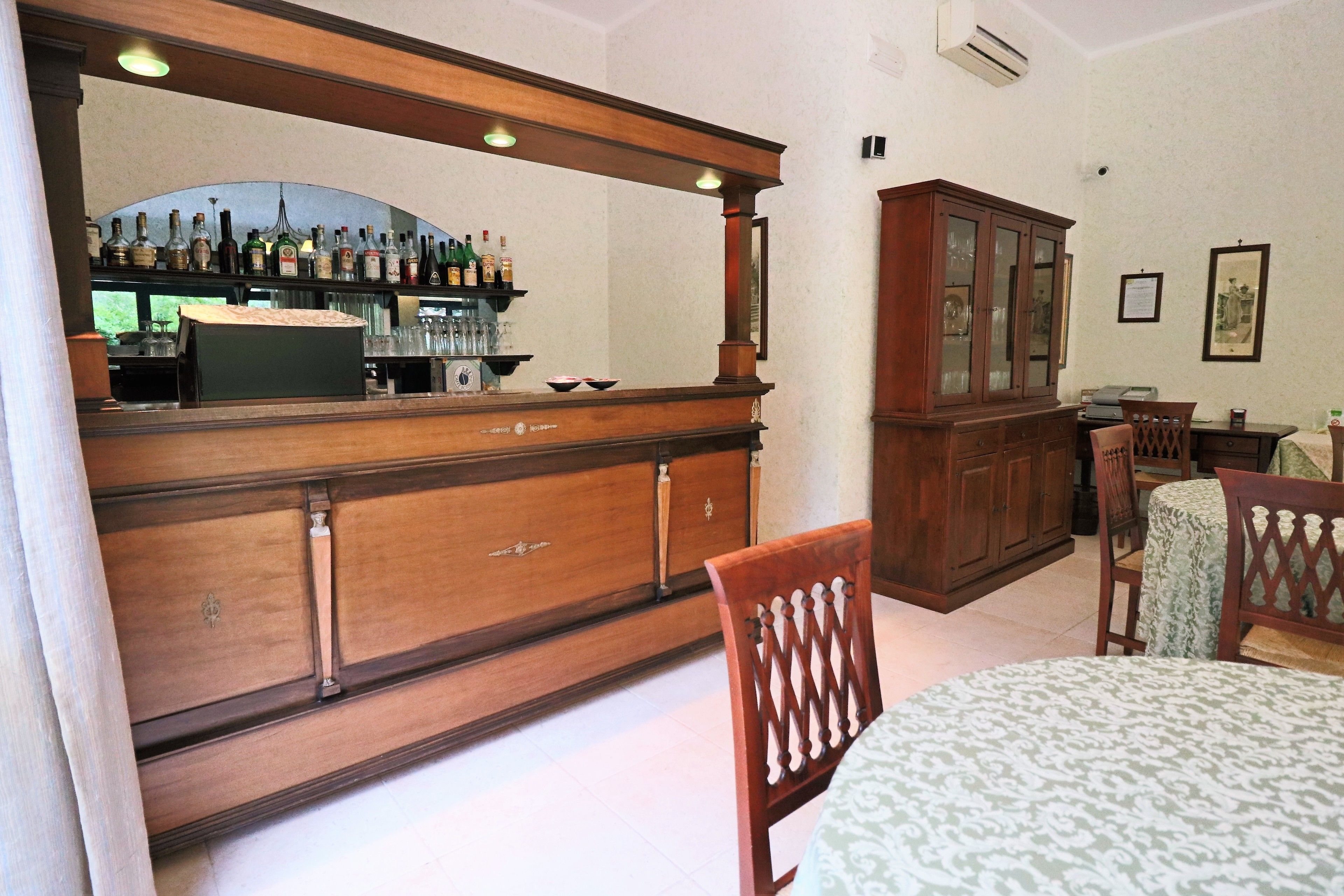 A bar area with a wooden counter, shelves with bottles, and a large mirror. There is a cabinet with glass doors and a dining table with chairs.