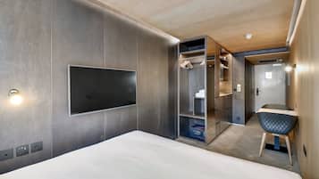 A modern hotel room with a large bed, a built-in TV, a wardrobe with hanging space, and a small seating area.