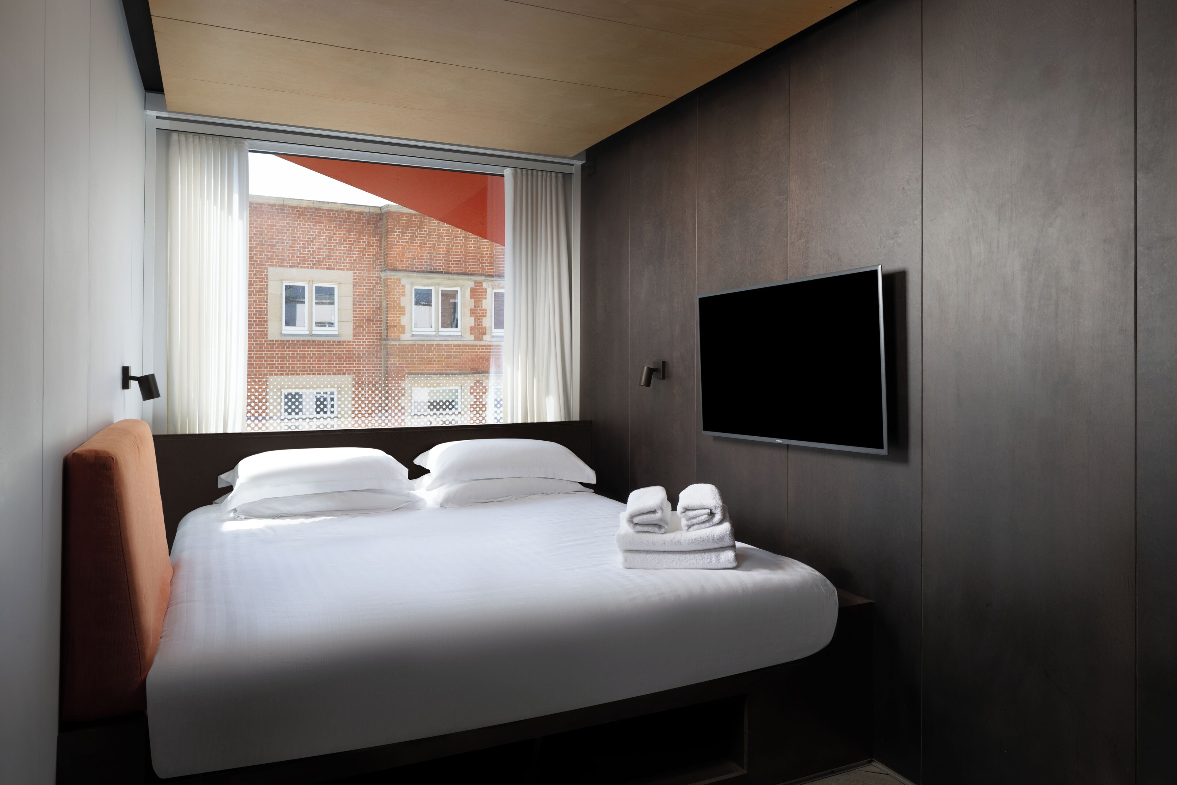 A modern hotel room with a large bed, a flat-screen TV mounted on the wall, and a window with curtains.