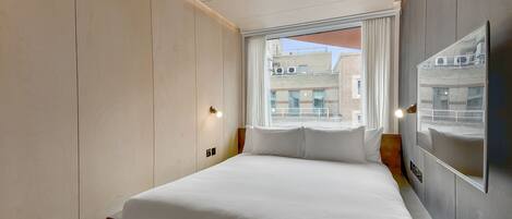 A modern hotel room with a large bed, a window with curtains, and a wall-mounted mirror.
