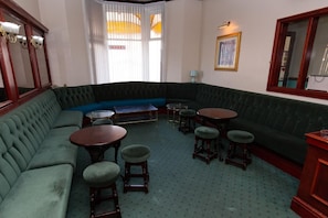 A corner room with a green upholstered sofa, a round wooden table, and matching stools. - The Riviera Hotel (Blackpool)