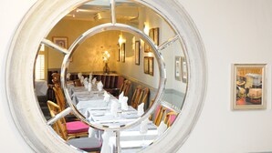 A dining area with a round mirror reflecting the room, wooden chairs, white tablecloths, and framed pictures on the wall. - The Dolphin Hotel (St. Martin)