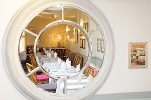 Un comedor con un espejo redondo que refleja la estancia, sillas de madera, manteles blancos y cuadros enmarcados en la pared.