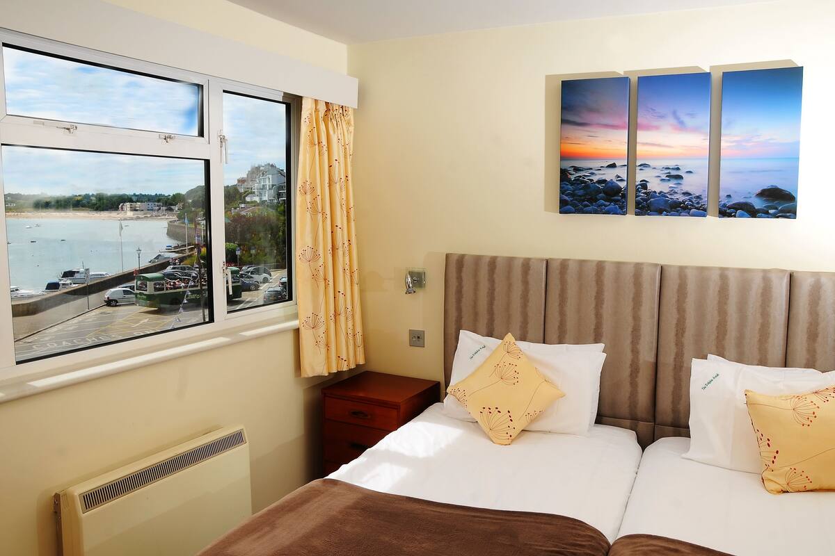 Habitación de hotel con cama, mesita de noche, ventana con vista al mar y barcos, y un cuadro de un atardecer.