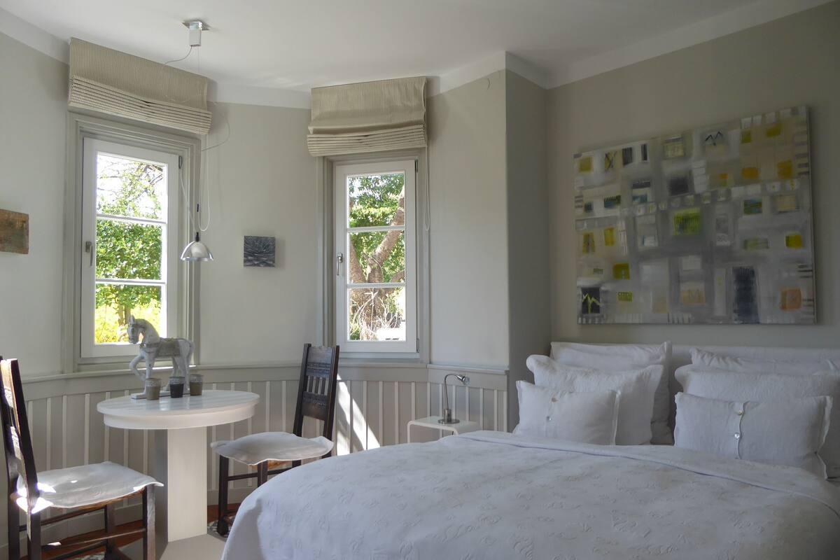 A bedroom with a large bed, two windows with curtains, a small table with a chair, and a wall with a bulletin board.