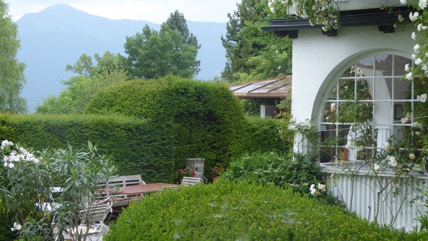 A lush garden with a white building featuring a large arched window. - Am Eichholz Galerie & Art Hotel (Murnau am Staffelsee)