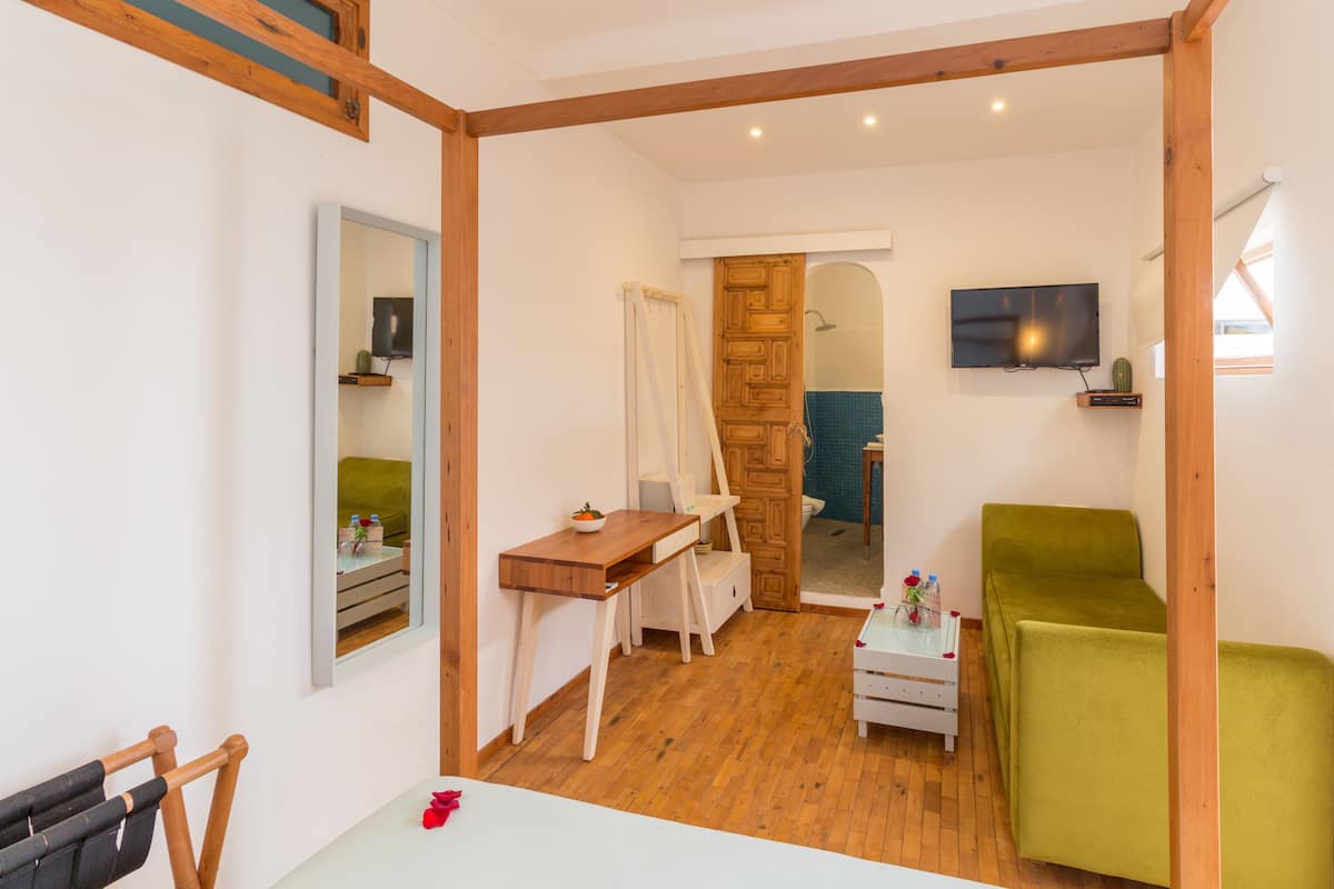 A room with a wooden floor, a green sofa, a wooden desk with a chair, a television mounted on the wall, and a wooden door leading to a bathroom.