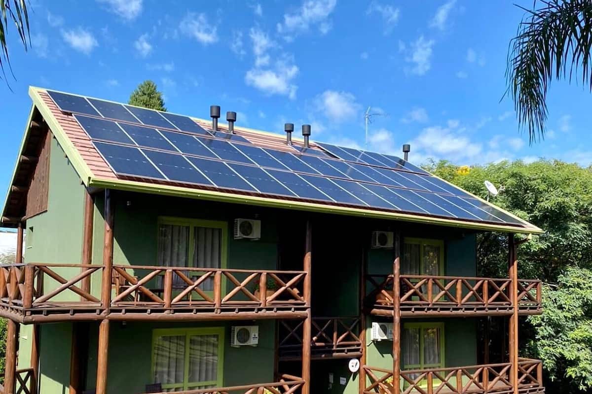 A two-story house with a sloped roof covered in solar panels, surrounded by lush greenery.