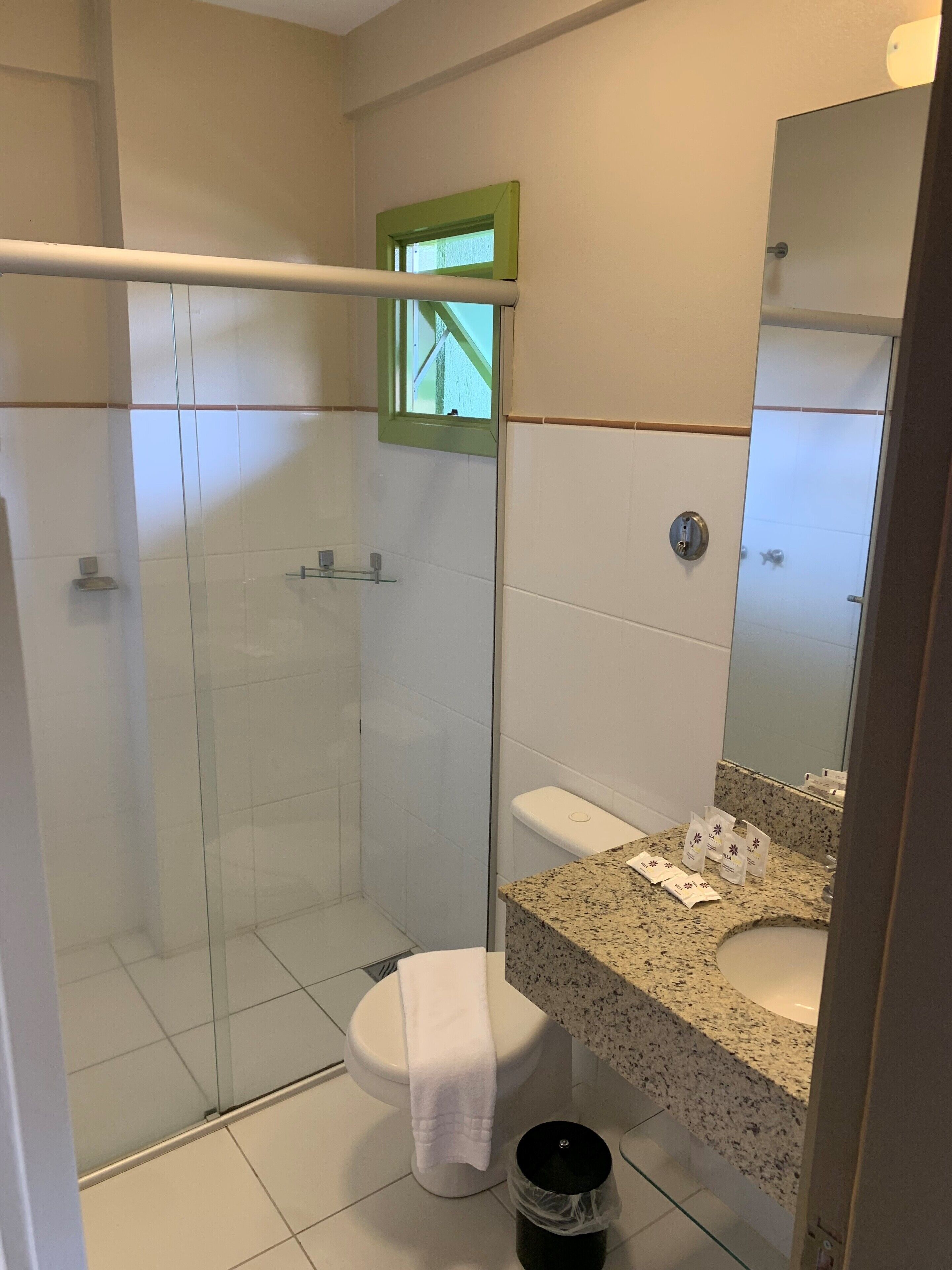 Une salle de bain dotée d’une douche à parois de verre, de toilettes blanches, d’un lavabo sur un plan de travail en granit et d’une petite fenêtre à cadre vert.
