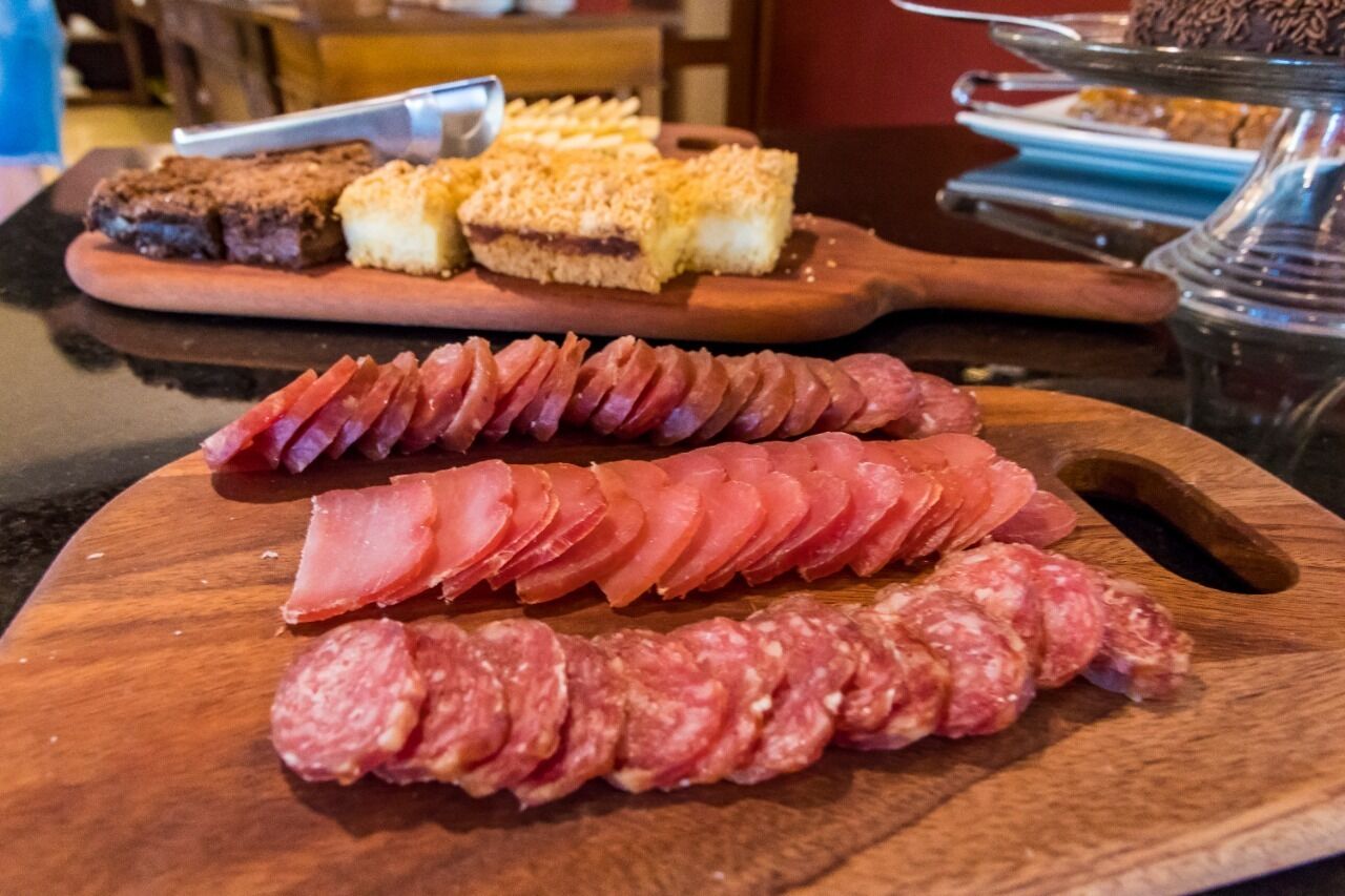 Una tabla de madera con embutidos y una bandeja de postres al fondo.