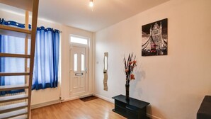 A room with a wooden floor, a door with a white frame, a blue curtain, a black console table with a vase, and a painting of a bridge. - Two Bedroom Serviced Apartment (Luton)