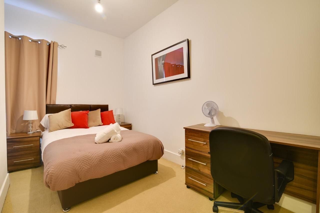 A hotel room with a bed, a desk, a chair, a nightstand, a lamp, a fan, and a picture on the wall.