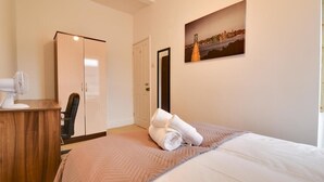 A hotel room with a bed, a wooden desk, a chair, a wardrobe, and a cityscape painting on the wall. - Two Bedroom Serviced Apartment (Luton)