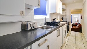A modern kitchen with white cabinets, a black countertop, a built-in oven, and a window with blue blinds. - Two Bedroom Serviced Apartment (Luton)