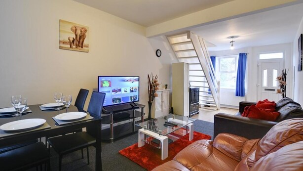 A modern living room with a dining area, a staircase, and a television displaying a menu. - Two Bedroom Serviced Apartment (Luton)