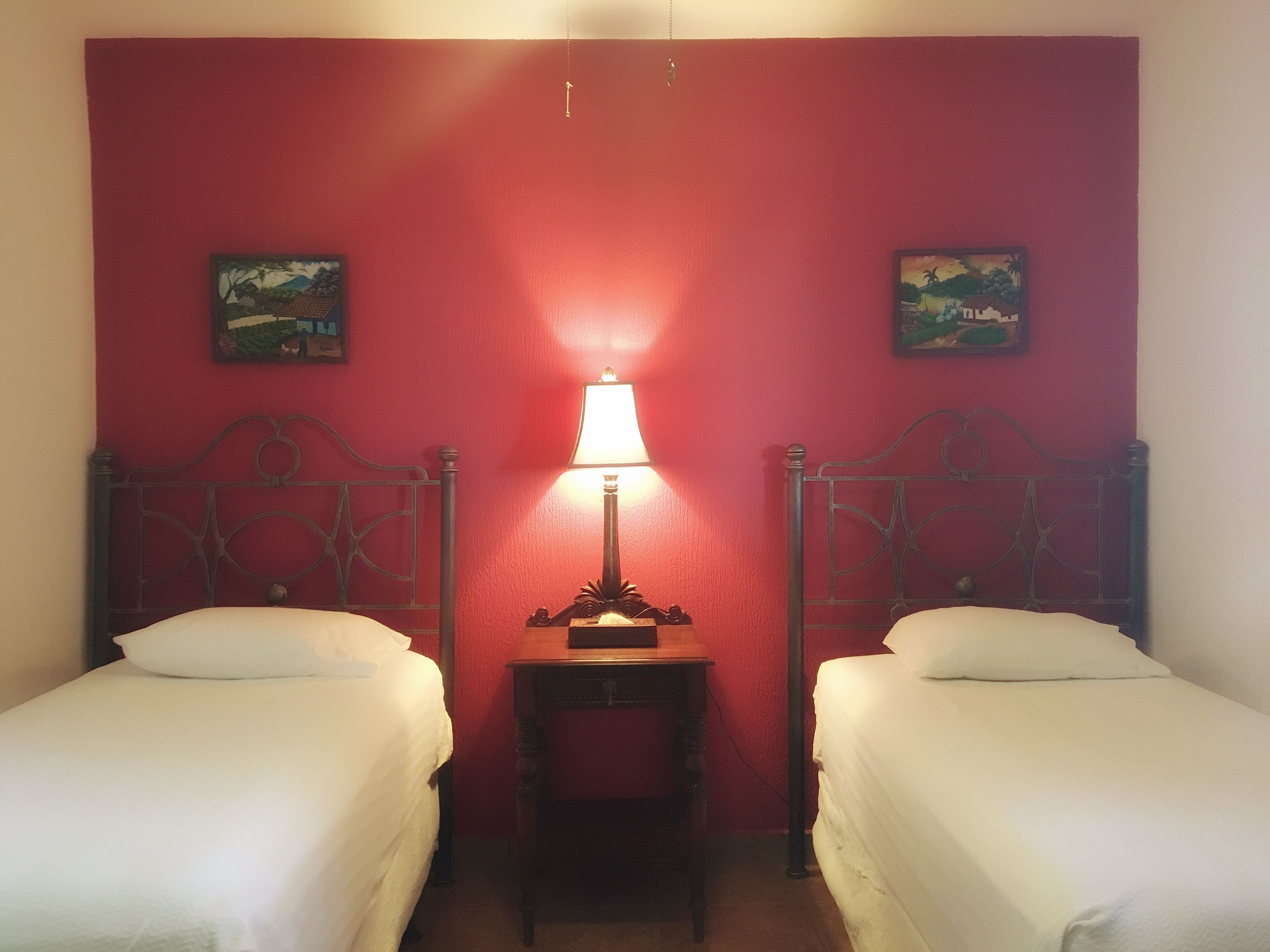 A room with two single beds, a nightstand with a lamp, and two wall-mounted paintings.