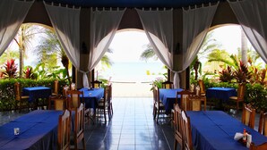 Un área de comedor al aire libre con mesas y sillas, vistas a la playa y plantas tropicales.