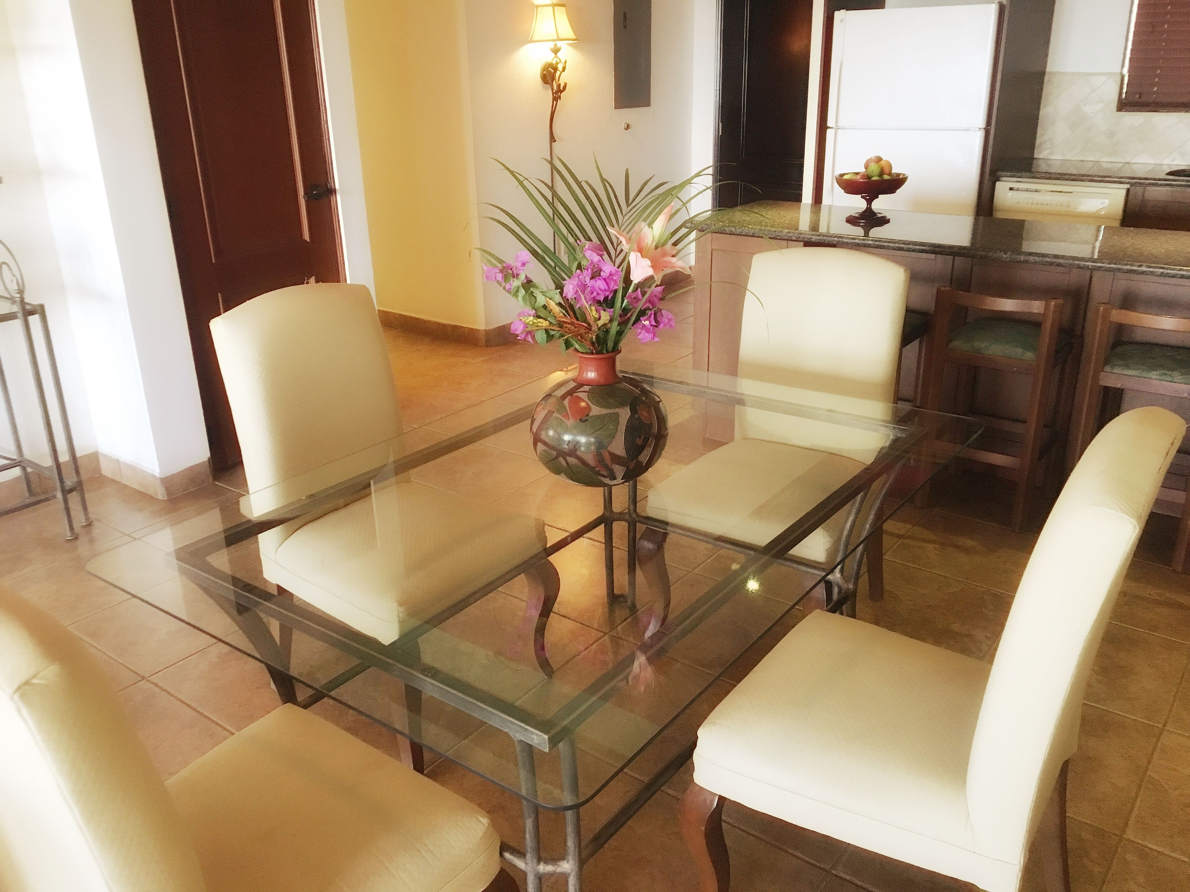 A dining area with a glass-top table surrounded by four chairs, a vase with flowers, and a kitchen in the background.