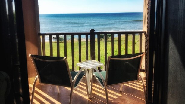 A balcony with two chairs and a small table, overlooking a grassy area and the ocean. - Suite San Marcos 124 Gran Pacifica Resort (Villa El Carmen)