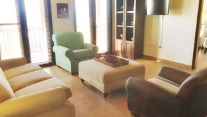 Una sala de estar con un sillón verde, sofás color beige y un sillón marrón. Hay un televisor de pantalla plana montado en la pared y un aparador con estantes.