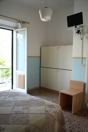 A bedroom with a bed, a wooden stool, a television mounted on the wall, and a window with curtains. - Hotel Ornella (Camaiore)