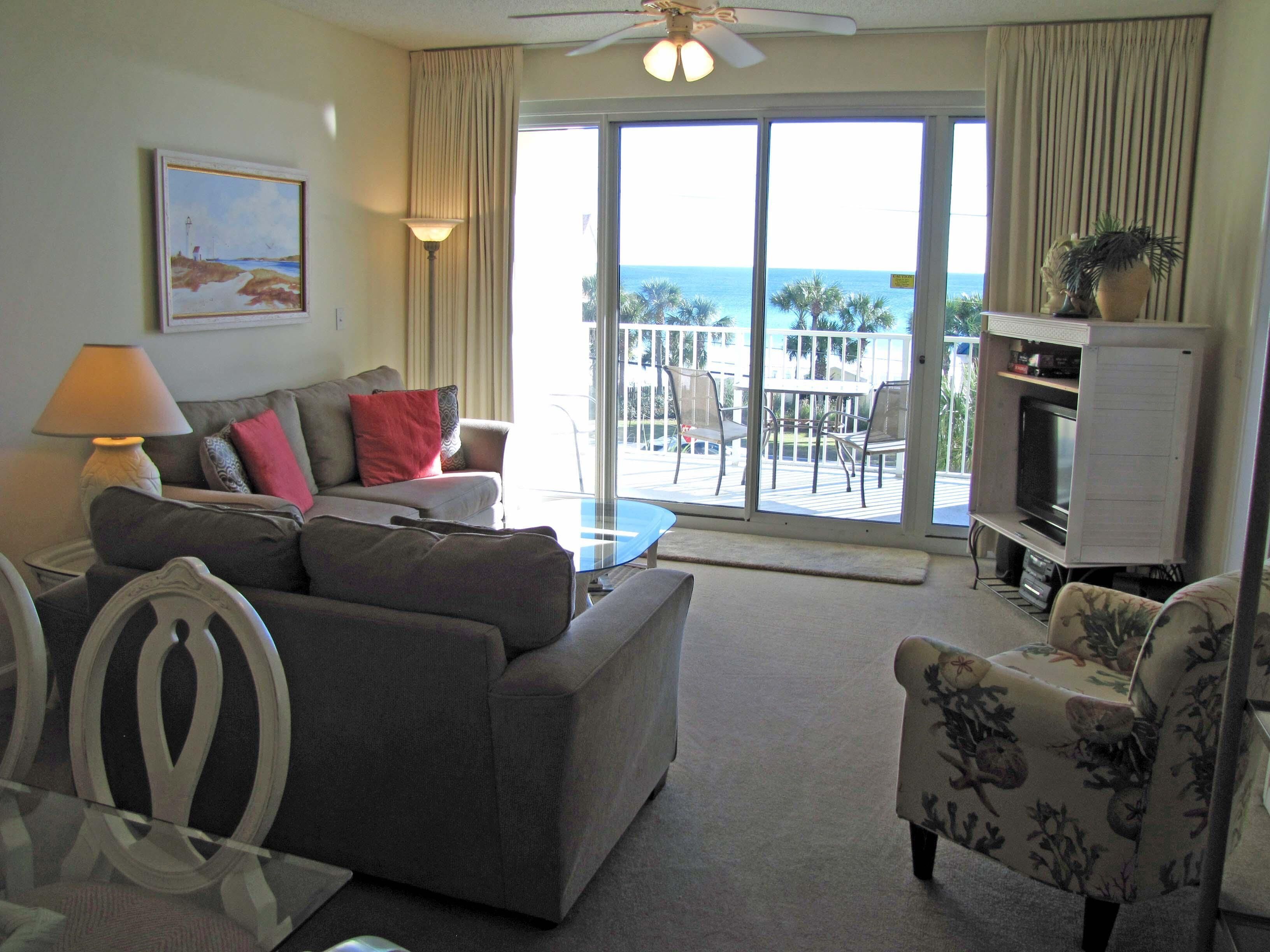 A living room with a sofa, armchair, and a TV stand. There is a balcony with a view of the ocean.