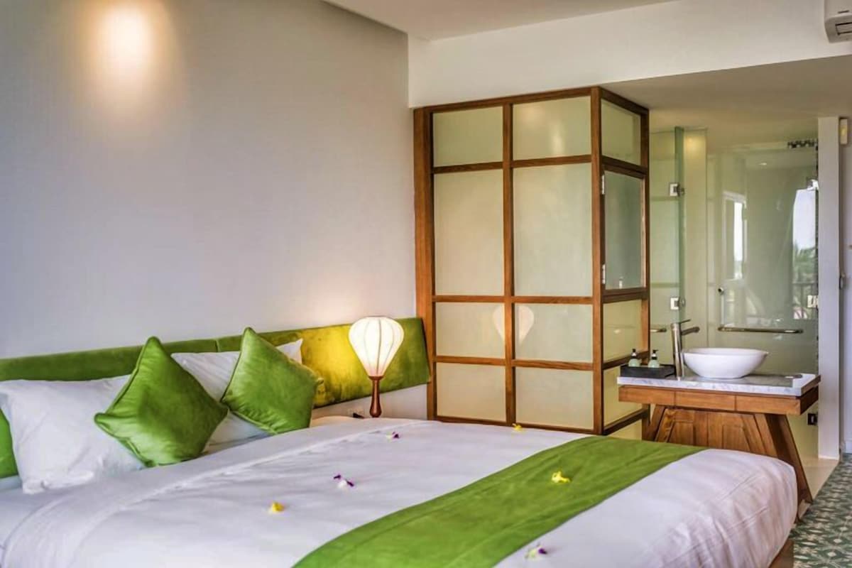 A hotel room with a bed, a green headboard, a bedside lamp, a large mirror, and a bathroom with a sink and mirror.