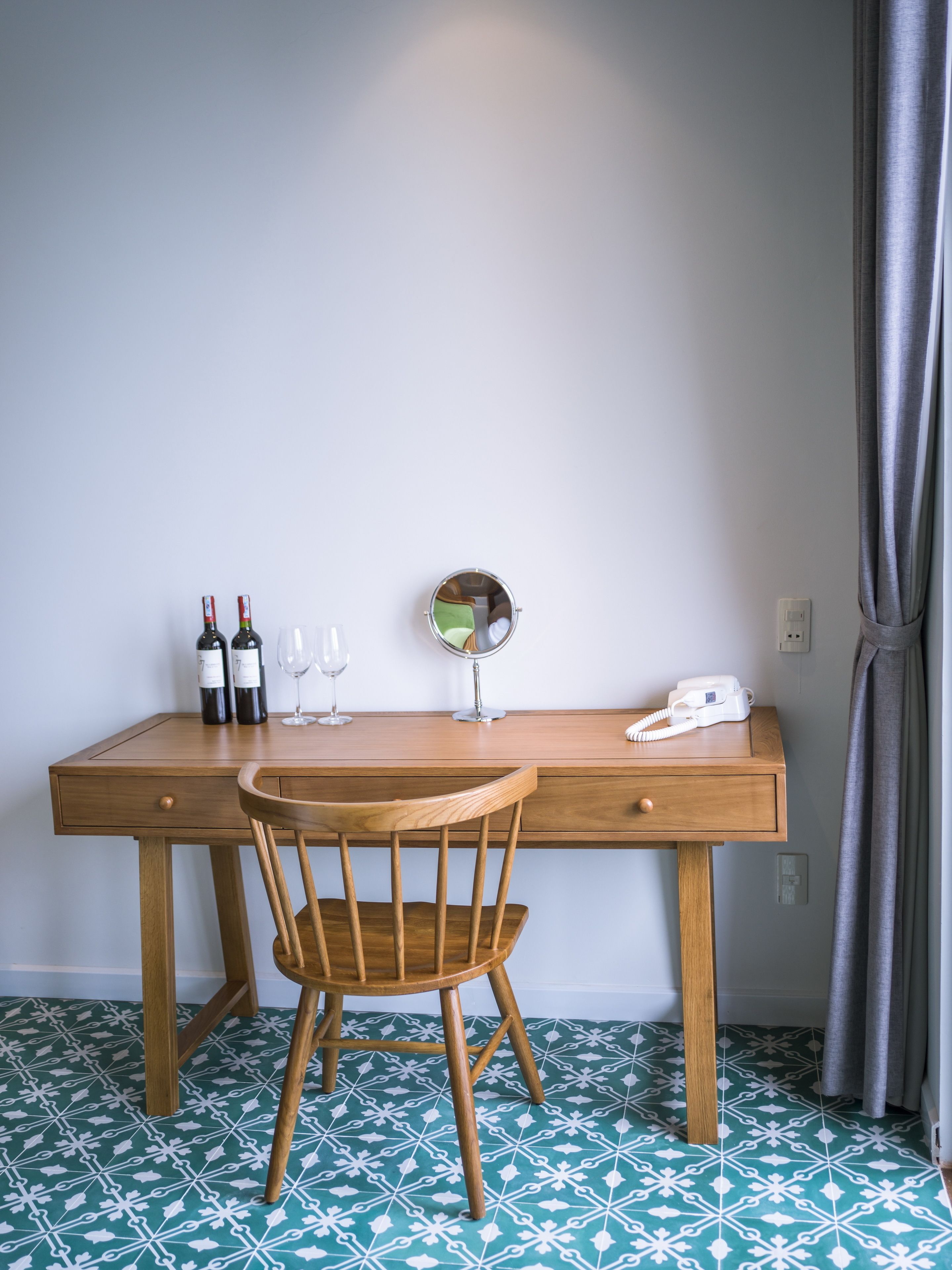 A wooden desk with a chair, a mirror, two wine bottles, and a telephone.