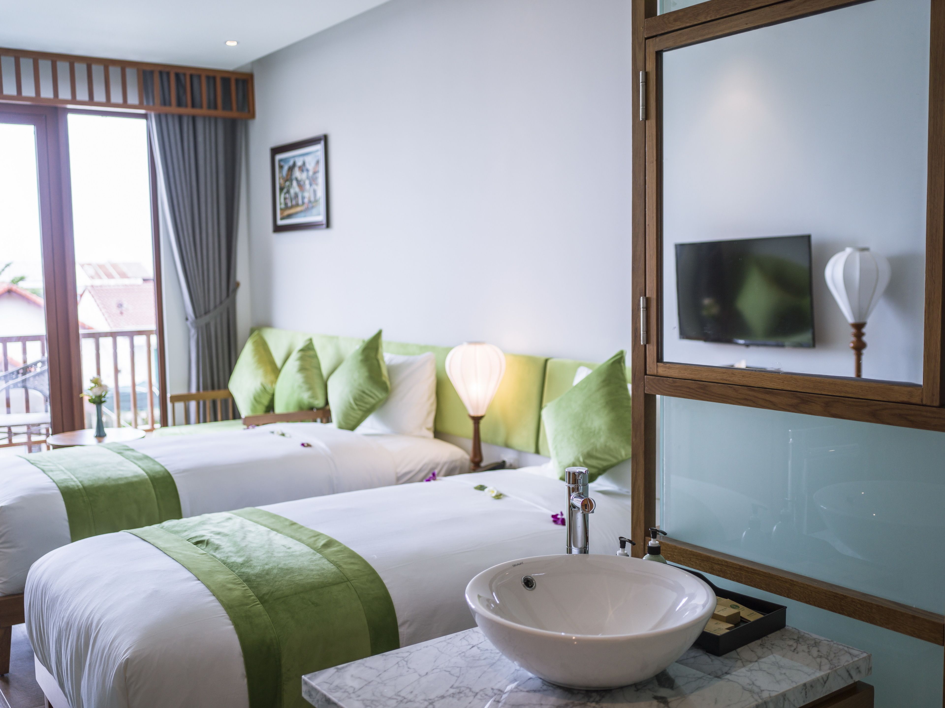 Habitación de hotel con dos camas, un espejo grande, un baño con lavamanos blanco y un balcón con vistas.