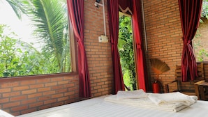 A room with a brick wall, a bed with white linens, red curtains, and a view of greenery outside. - Gibbon Singing Homestay (Nam Cat Tien)