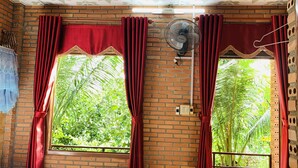 A room with brick walls, two windows with red curtains, a ceiling fan, and a view of greenery outside. - Gibbon Singing Homestay (Nam Cat Tien)