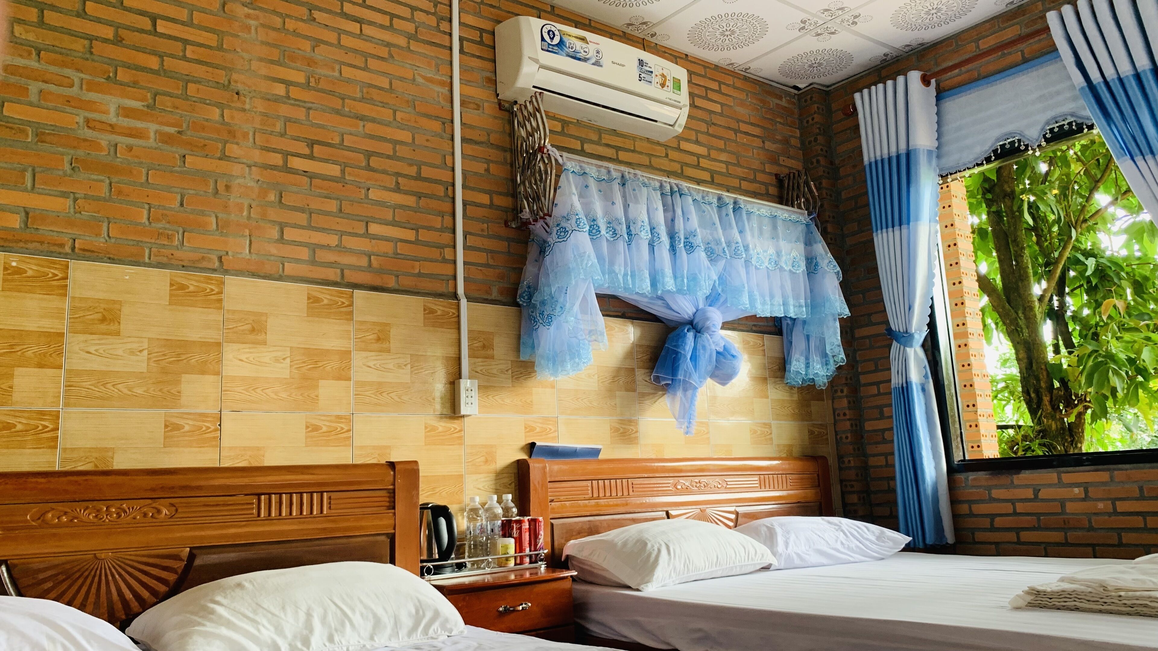 A hotel room with a brick wall, a wooden headboard, a bed with white linens, a nightstand with a kettle, and a window with blue curtains.