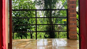A room with a tiled floor, a brick wall with holes, and a view of lush green foliage through a window with metal bars. - Gibbon Singing Homestay (Nam Cat Tien)