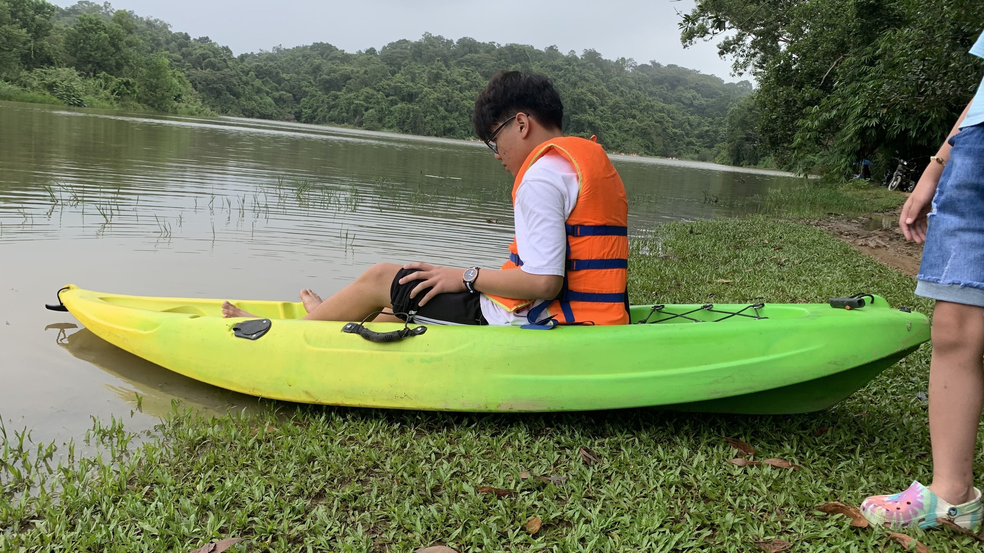 Une personne est assise sur un kayak vert, près d’un lac paisible, et porte un gilet de sauvetage orange.