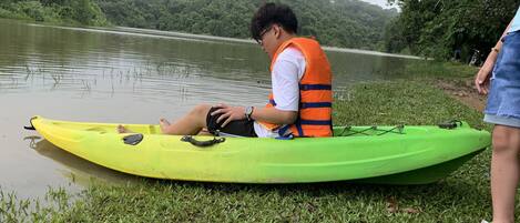 Une personne est assise sur un kayak vert, près d’un lac paisible, et porte un gilet de sauvetage orange.